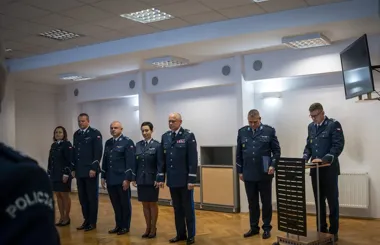 Funkcjonariusze policji w mundurach stoją w sali podczas uroczystego spotkania, a jeden z nich przemawia zza pulpitu.