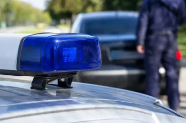 Policyjny radiowóz z niebieskim światłem ostrzegawczym na dachu, w tle funkcjonariusz i inny samochód.